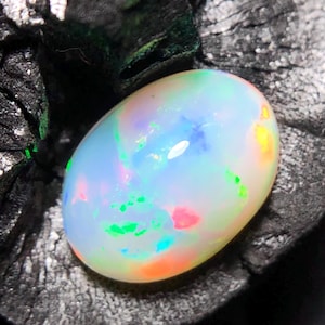 Könnte beinhalten: Ein ovales Opal-Edelstein mit milchig-weißer Grundfarbe. Der Opal zeigt lebhafte Farbblitze, darunter Grün, Blau, Rosa und Orange, wodurch ein Farbspiel-Effekt entsteht. Der Edelstein ist vor einem dunklen, strukturierten Hintergrund platziert.