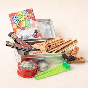 May include: A silver metal box filled with smoking accessories. Items include rolling papers, a grinder, a container, and two green tubes. A package of "Zkittlez" candy is also visible.
