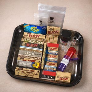 Puede incluir: Una bandeja negra llena de accesorios para fumar. Incluye paquetes de papel de liar de marcas como RAW y OCB, junto con un molinillo, recipientes y un recipiente rojo y morado etiquetado como "Cannabis".