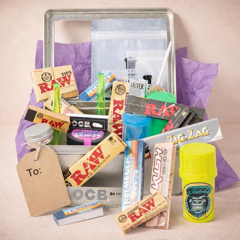 Puede incluir: Una caja met&aacute;lica llena de accesorios para fumar, incluyendo papel de liar, filtros y un molinillo. Los art&iacute;culos est&aacute;n dispuestos sobre papel de seda morado. Varias marcas son visibles, incluyendo RAW, OCB y Zig-Zag.