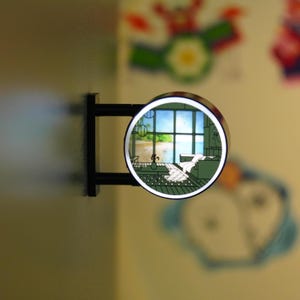Puede incluir: Un letrero redondo e iluminado con un soporte negro. El letrero presenta una ilustración de dibujos animados de una habitación con vista a una playa y al océano. Está montado en una pared de color claro.