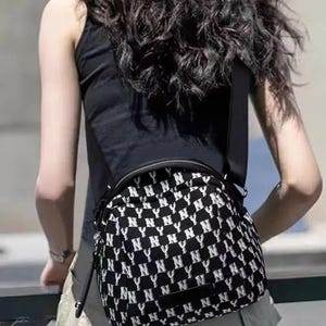 May include: Black and white crossbody bag with a repeating "NY" pattern. The bag has a black shoulder strap and a zippered top. The bag is worn over the shoulder.