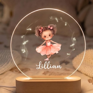 May include: A circular acrylic night light featuring an illustration of a young ballerina in a pink tutu with the name Lillian printed below. The light is mounted on a smooth, oval-shaped wooden base and emits a warm glow, surrounded by musical notes.