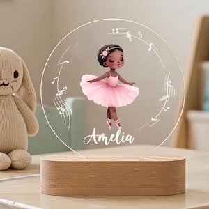 Op de afbeelding: Een rond acryl nachtlampje met een gedrukte illustratie van een ballerina in een roze tutu, de naam Amelia en muzieknoten. De doorzichtige schijf is gemonteerd op een ovale houten voet met een stroomkabel aan de zijkant.