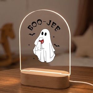 Puede incluir: Una luz nocturna de acrílico arqueada con base de madera que presenta una ilustración de un fantasma blanco sosteniendo una copa de vino tinto. El texto BOO-JEE está impreso en negro sobre el fantasma, junto a pequeños iconos de murciélagos.