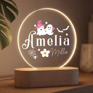 May include: A circular acrylic night light with a warm glow, featuring the name Amelia Miller in white text. The design includes two white ghosts, one wearing a pink witch hat, a spider web, a spider, a white flower, and two small black bats.