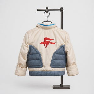 Könnte beinhalten: Eine beige-blaue Jacke mit einem roten Fuchsemblem auf der Rückseite. Die Jacke hat einen hohen Kragen mit blauem Rand, lange Ärmel mit breiten Bündchen sowie gesteppte blaue Einsätze am unteren Rücken und an den Seiten.