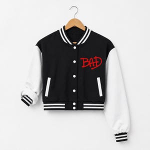 May include: Black and white varsity jacket with the word "BAD" in red script on the chest. The jacket has white sleeves, a black body, and white and black striped trim on the collar, cuffs, and hem. It is buttoned up and hanging on a wooden hanger.