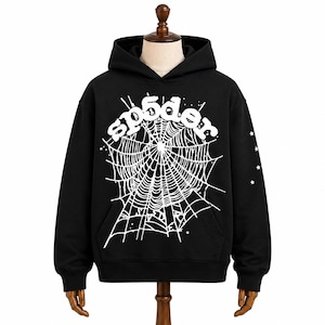 May include: A black hooded sweatshirt featuring a large white graphic on the front. The graphic displays a spider web design with the text sp5der arched above it. Small white stars are scattered around the web and on the right sleeve.