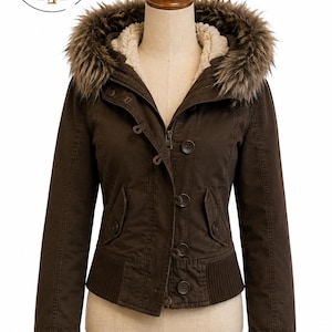Könnte beinhalten: Eine dunkelbraune Jacke mit einer Kapuze mit Kunstfellbesatz, einem cremefarbenen Sherpa-Futter, einem Frontreißverschluss und einer Knopfleiste. Die Jacke verfügt über zwei Seitentaschen mit Knopfklappen und einen gerippten Saum an der Taille.