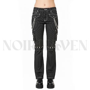 Könnte beinhalten: Schwarze Nadelstreifenhose mit weißen Kontrastnähten, silberfarbenen Metallketten an den Vordertaschen, seitlichen Cargotaschen und einem horizontalen Band aus silbernen Nieten über jedem Knie, mit Knopf- und Reißverschluss.
