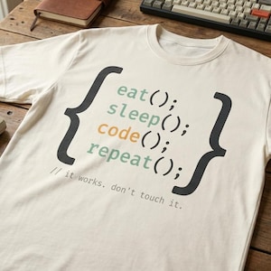 May include: Cream-coloured t-shirt featuring the text "eat(); sleep(); code(); repeat(); // it works. don't touch it." in black, green, and orange. The shirt is on a wooden table with keyboards and a coffee cup.