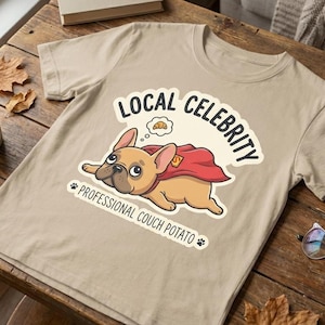 Puede incluir: Camiseta color beige con un dibujo de un bulldog francés de dibujos animados con una capa roja y las palabras "LOCAL CELEBRITY" y "PROFESSIONAL COUCH POTATO". La camiseta está sobre una mesa de madera con una taza de café y gafas.