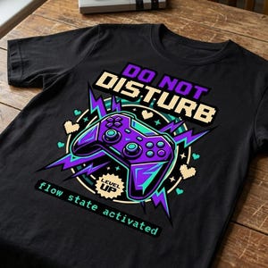 May include: Black t-shirt with a video game controller graphic in purple, teal, and white. Text on the shirt reads "DO NOT DISTURB" and "flow state activated". A retro game controller and headphones are visible.