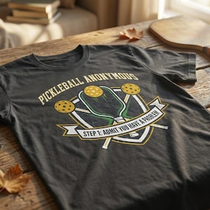 Può includere: T-shirt grigio scuro con la scritta "PICKLEBALL ANONYMOUS" e una grafica di racchette e palline da pickleball. La maglietta ha anche la scritta "STEP 1: ADMIT YOU HAVE A PROBLEM". La maglietta è su un tavolo di legno.