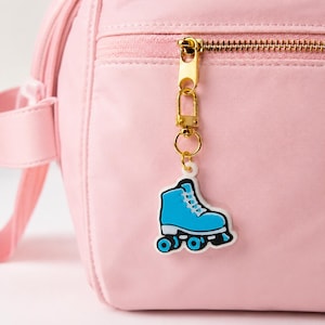 Könnte beinhalten: Ein hellrosa Rucksack mit goldenem Reißverschluss und einem blauen Rollschuh-Anhänger. Der Anhänger ist im Cartoon-Stil mit weißer Umrandung und schwarzen Rollen gestaltet. Der Rucksack besteht aus glattem Material.