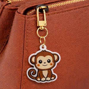 Op de afbeelding: Een bruine en witte apenbedel met een goudkleurige sluiting, bevestigd aan een bruine leren tas. De bedel heeft een lachend apenontwerp met een witte omtrek.