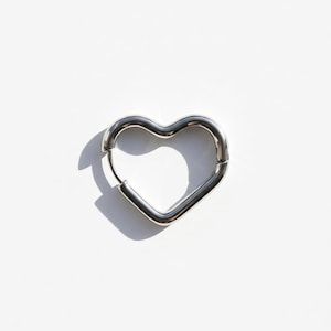Op de afbeelding: Een enkele, zilverkleurige oorbel in de vorm van een hart. De oorbel is gemaakt van glad, glanzend metaal en heeft een eenvoudig, minimalistisch ontwerp. De oorbel wordt getoond tegen een witte achtergrond, met een schaduw naar links.