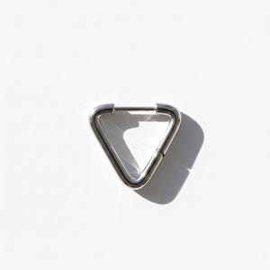 Op de afbeelding: Een enkele, glanzende zilveren oorbel in een driehoekige vorm. De oorbel heeft een scharniersluiting en staat tegen een helderwitte achtergrond, waardoor een schaduw ontstaat. De sieraden zijn minimalistisch en modern.
