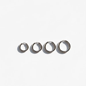 Peut inclure: Quatre boucles d'oreilles créoles argentées de tailles différentes sont disposées en ligne sur un fond blanc. Les boucles d'oreilles sont circulaires et présentent un design simple et minimaliste. L'éclairage crée des ombres.