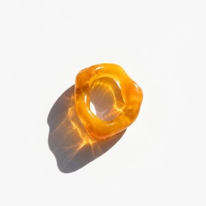 May include: A translucent, amber-colored ring with an irregular, organic shape. The ring is made of a glossy material and casts a shadow on the white surface below. The ring's design is modern and minimalist.