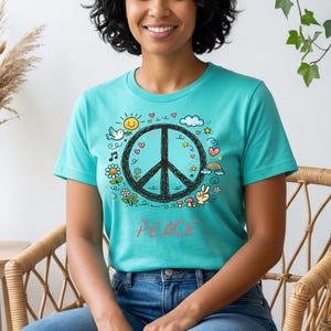Könnte beinhalten: Ein türkisfarbenes T-Shirt mit einem schwarzen Friedenszeichen, umgeben von bunten Kritzeleien, darunter eine Sonne, Herzen und ein Regenbogen. Das Wort "PEACE" steht in roter Schrift darunter. Das T-Shirt wird von einer Person getragen.