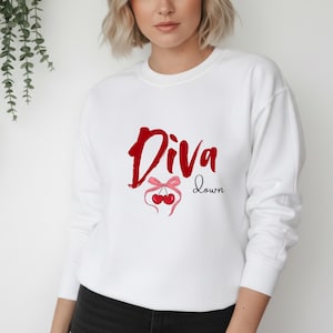 Könnte beinhalten: Weißes Sweatshirt mit dem Text "Diva Down" in roter Schrift. Darunter eine rosa Schleife und zwei rote Kirschen. Das Sweatshirt hat einen Rundhalsausschnitt und lange Ärmel.