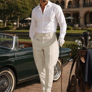 May include: A white long-sleeved button-down shirt with a pleated front panel paired with high-waisted cream-colored trousers. The outfit includes black leather tassel loafers and a white golf glove, with a golf club held in one hand.