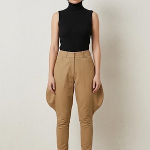 May include: A sleeveless black ribbed turtleneck top paired with tan jodhpurs featuring flared hip panels, a structured waistband with belt loops, a front zipper fly, and tapered legs that extend down to the ankles, worn with neutral-toned pumps.