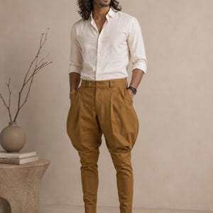 May include: A long-sleeved, off-white button-down shirt with a collar, paired with tan, high-waisted trousers featuring a pleated, voluminous hip design that tapers toward the ankles. The outfit is completed with tan suede loafers and a dark wristwatch.