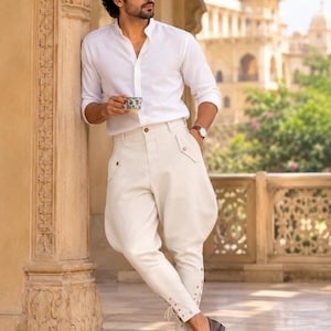 May include: A white long-sleeved button-down shirt with a mandarin collar paired with off-white jodhpurs featuring side pockets, button details, and lace-up closures at the ankles. The outfit is completed with dark brown leather tassel loafers.
