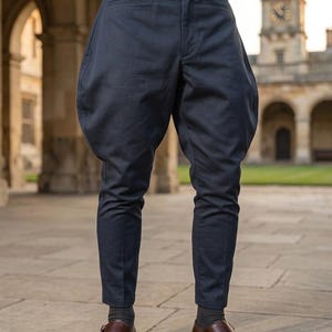 May include: Dark navy blue jodhpurs featuring a flared hip design, a fitted lower leg, and a high waist. The trousers are paired with a brown leather belt with a gold-toned buckle, dark grey socks, and brown leather double monk strap shoes.