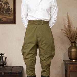 May include: A crisp white long-sleeved button-down shirt paired with olive green high-waisted breeches featuring a wide, flared hip design and a buttoned waistband, worn with tall black leather boots that reach just below the knee.