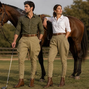 May include: Two individuals wearing khaki breeches with lace-up ankles, brown leather belts, and brown leather boots. One wears an olive green button-down shirt, and the other wears a white button-down shirt. Both stand beside a brown horse.