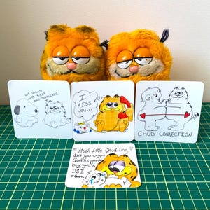 May include: Two plush Garfield toys sit above four illustrated cards featuring Garfield and other characters. The cards have text that reads "We should get beer and dumplings," "I miss you," "Chud Connection," and "Hush little Chudling, don't you cry..."