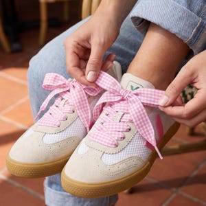 May include: A pair of off-white sneakers with tan soles and pink accents. The sneakers feature white mesh panels and are laced with pink and white gingham ribbon. The shoes are being tied with the ribbon.