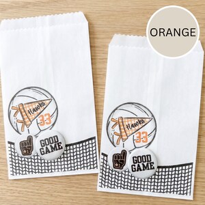 May include: Two white paper treat bags with volleyball-themed graphics. Each bag features a volleyball illustration, a pennant that says "Hawks 33", and two buttons that say "Good Game" and a foam finger. A black net design is at the bottom.