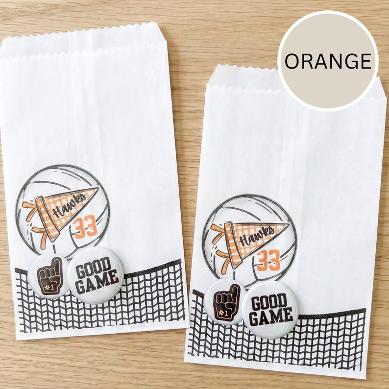 May include: Two white paper treat bags with volleyball-themed graphics. Each bag features a volleyball illustration, a pennant that says "Hawks 33", and two buttons that say "Good Game" and a foam finger. A black net design is at the bottom.