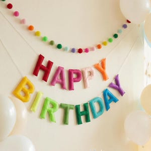 May include: A colorful felt banner spelling "HAPPY BIRTHDAY" in various colors, with a string of rainbow-colored felt balls above. White balloons are visible in the background, creating a festive party atmosphere.