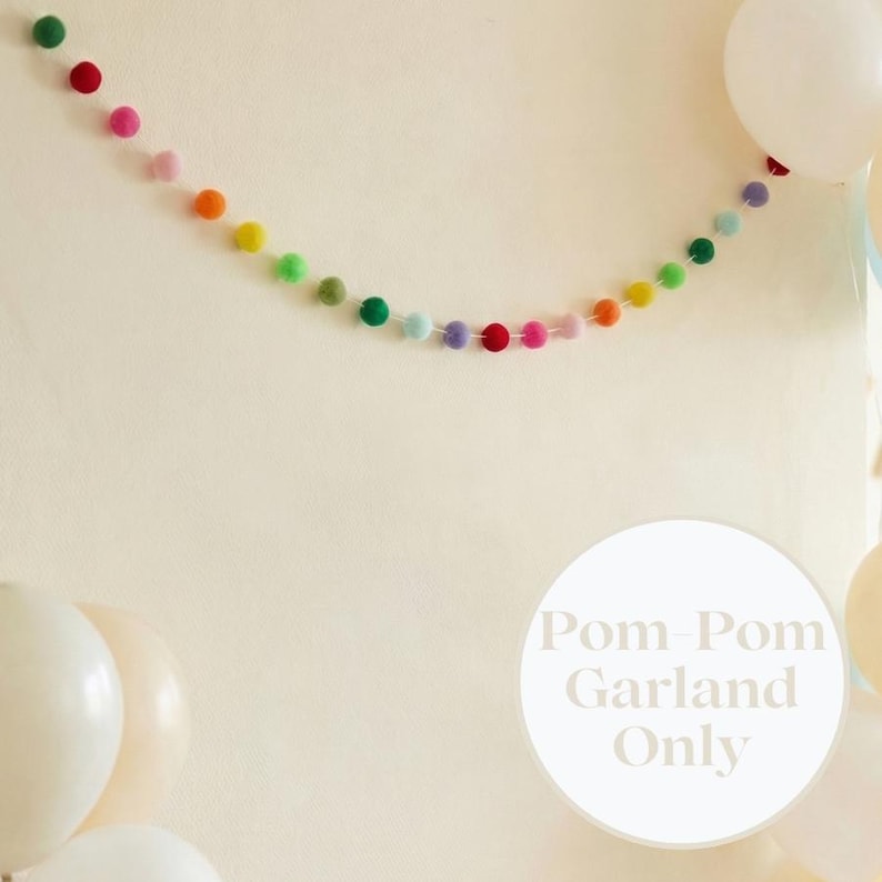 May include: A vibrant pom-pom garland in a spectrum of colours, including red, pink, orange, yellow, green, and blue. The garland is set against a cream backdrop, with white balloons in the foreground. The text "Pom-Pom Garland Only" is in a white circle.