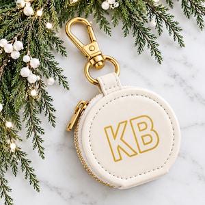 Puede incluir: Un estuche redondo de color blanco roto con las letras KB en color dorado grabadas en la parte frontal. El estuche incluye una cremallera de metal dorado y un cierre de mosquetón a juego en la parte superior para colgarlo fácilmente.