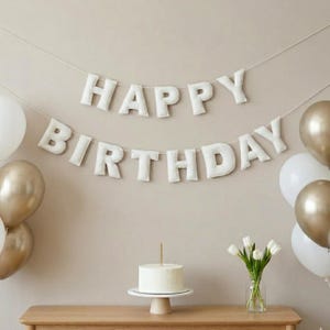 May include: A birthday party scene with white and gold balloons, a cake, and a banner that says "HAPPY BIRTHDAY." The banner is made of white fabric letters strung on a string. A small white cake sits on a white cake stand with a single gold candle.