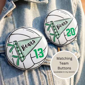 May include: Two white and black volleyball-themed buttons with the word "Bears" on a green pennant. One button has the number 13, the other has 20. The buttons are on a denim jacket.