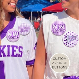 Puede incluir: Dos personas vestidas con ropa blanca y morada de los Northwest Huskies. La imagen presenta varios botones redondos con el logotipo y el texto del equipo. Los botones tienen 5,7 cm de diámetro. El fondo incluye una carpa roja y azul.