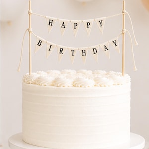 May include: A white frosted cake topped with two tiers of cream-coloured fabric pennant banners suspended between two wooden sticks. The top banner spells HAPPY and the bottom banner spells BIRTHDAY in black capital letters on triangular flags.