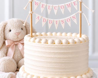 May include: A round white frosted cake topped with two wooden dowels holding two rows of triangular pennant flags. The top row spells WELCOME in pink letters, and the bottom row spells ADDISON in pink letters. A plush beige bunny sits nearby.