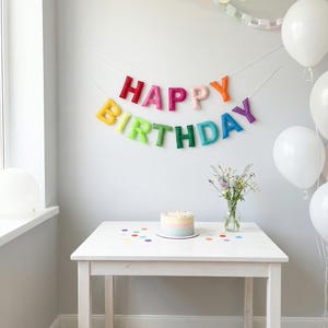 May include: A birthday party setting featuring a white table, a rainbow cake, and a colourful "HAPPY BIRTHDAY" banner. White balloons and confetti enhance the celebratory feel. A vase of flowers is on the table.