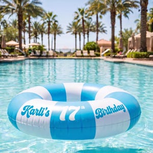 May include: A round inflatable pool float featuring alternating light blue and white stripes. The text Karli's 17th Birthday is printed in a bold, blue, retro-style font across the front of the float, which is resting on the surface of a swimming pool.