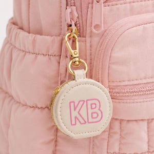 Könnte beinhalten: Ein altrosa gesteppter Rucksack mit einem goldfarbenen Karabinerhaken, an dem ein kleines, rundes, cremefarbenes Reißverschlusstäschchen befestigt ist. Das Täschchen ist mit den Buchstaben KB in leuchtend rosa Umrandung versehen.