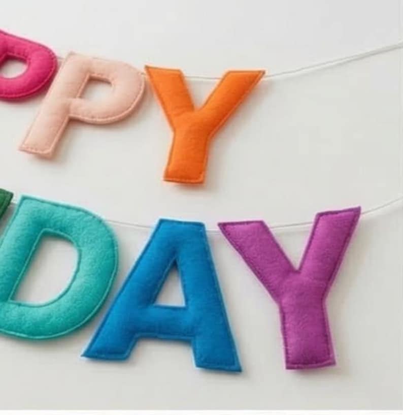 May include: A colourful felt banner spelling "HAPPY DAY". The letters are in various colours, including pink, peach, orange, teal, blue, and purple. The letters are strung together with a white string, ideal for a birthday celebration.